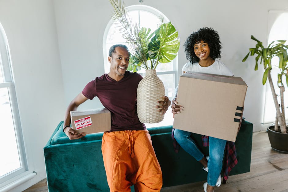 A man and a woman inside a bright, modern home are preparing for a house move, holding cardboard boxes and a large ceramic vase with green leafy plants. The man is wearing an orange pair of trousers a