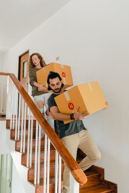 A professional mover from Strand Removals wearing a navy-blue work uniform and a black bandana with white patterns, standing inside a residential property next to a cardboard box labeled 'Class' and s
